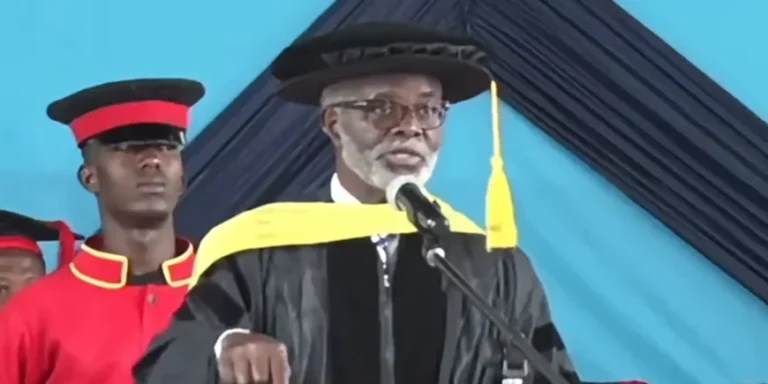 Elderly man in a black graduation gown and cap speaks at a podium, yellow stole draped over shoulders, microphone in front.