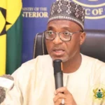 Man wearing a patterned cap and glasses speaks into a microphone at a press briefing, with a Ministry of Interior backdrop behind him.