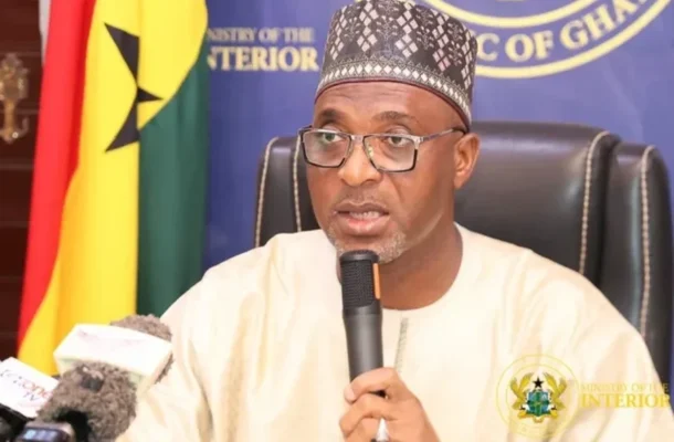 image_870x_6527f52779219 | Ghanaguardian.com Man wearing a patterned cap and glasses speaks into a microphone at a press briefing, with a Ministry of Interior backdrop behind him.
