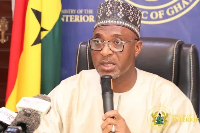 Man wearing a patterned cap and glasses speaks into a microphone at a press briefing, with a Ministry of Interior backdrop behind him.