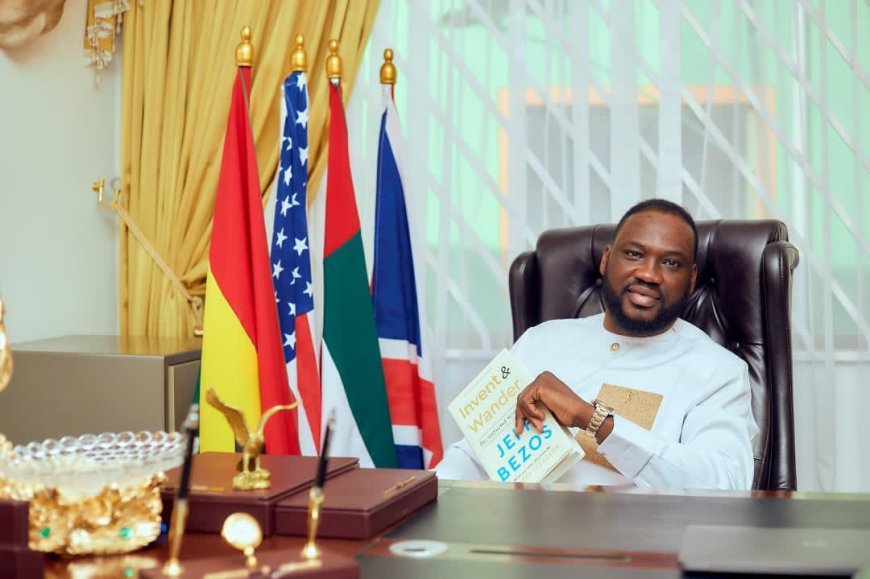 A man in a light-colored traditional outfit sits in a leather office chair behind a desk, holding a book with flags in the background. flags of several countries stand behind him along with gold desk ornaments and a window with blinds.