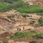 Workers dig in a muddy excavation site with small water-filled pools and a wooden plank bridge nearby, surrounded by green vegetation.