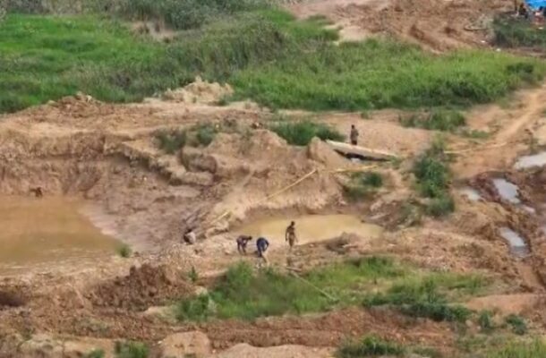 image_870x_6a1992048a364 | Ghanaguardian.com Workers dig in a muddy excavation site with small water-filled pools and a wooden plank bridge nearby, surrounded by green vegetation.