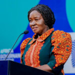 Woman speaker at a podium with microphone, wearing a red patterned blouse and green vest, at a blue backdrop.