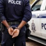 Uniformed police officer with hands cuffed behind back standing beside a police car