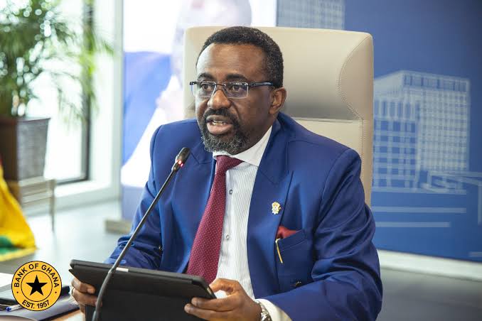 A man in a blue suit and red tie speaking into a microphone while holding a tablet, with a Bank of Ghana emblem visible on the desk seal nearby.