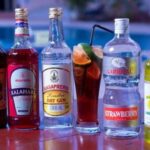 Row of colorful liquor bottles lined up on a poolside table with a glass of mixed drink nearby.