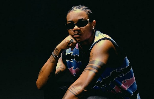 Portrait of a man wearing dark sunglasses and a colorful sleeveless vest, resting his chin on his hand in a dark backdrop.
