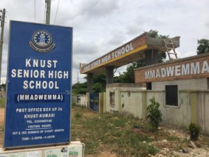 Blue signboard reads 'KNUST SENIOR HIGH SCHOOL (MMADWEMMA)' with address and hours, standing near the entrance gate.