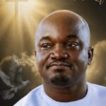 Close-up of a smiling Black man with a shaved head wearing a white shirt, with a dove and sunlit clouds in the background.