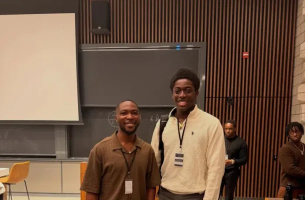 Two men stand in a classroom posing for a photo, wearing lanyards and smiles, with a whiteboard and wooden panel backdrop behind them.
