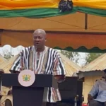 A man in a striped shirt speaks at a podium with a seal, under a colorful ceremonial tent, flanked by officials and soldiers.