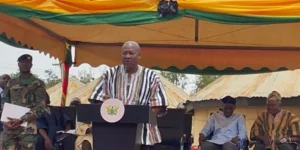 A man in a striped shirt speaks at a podium with a seal, under a colorful ceremonial tent, flanked by officials and soldiers.