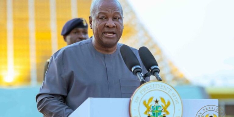 Older man speaks at a lectern with two microphones, wearing a gray traditional outfit; a security guard stands in the background outdoors.