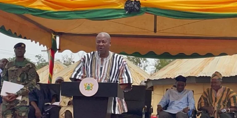 A man in a striped shirt speaks at a podium with a seal, under a colorful ceremonial tent, flanked by officials and soldiers.