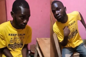 Two men with dark skin wearing matching yellow t‑shirts that say “KEEP CALM” pose in a pink-walled room; one sits at a wooden table looking down, the other sits sideways and points to himself with a serious expression.