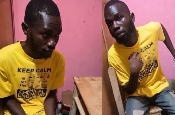 Two men with dark skin wearing matching yellow t‑shirts that say “KEEP CALM” pose in a pink-walled room; one sits at a wooden table looking down, the other sits sideways and points to himself with a serious expression.