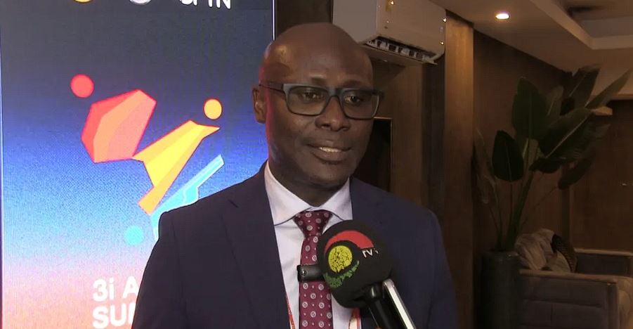 Man in a navy suit and red tie being interviewed, holding a microphone with a TV network logo visible.