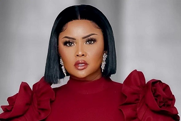 Close-up of a woman with a straight black bob, bold makeup, wearing a high-neck red dress with large ruffle sleeves and dangling diamond earrings.