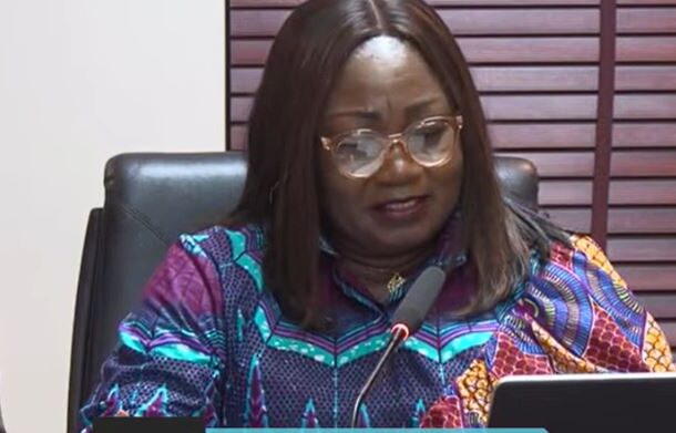 Woman in a colorful patterned blouse speaks into a microphone at a panel, with a tablet on the table beside her.