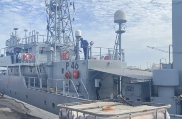 Grey patrol/research vessel docked at a pier, with crew on the upper deck near antennas and radar domes.