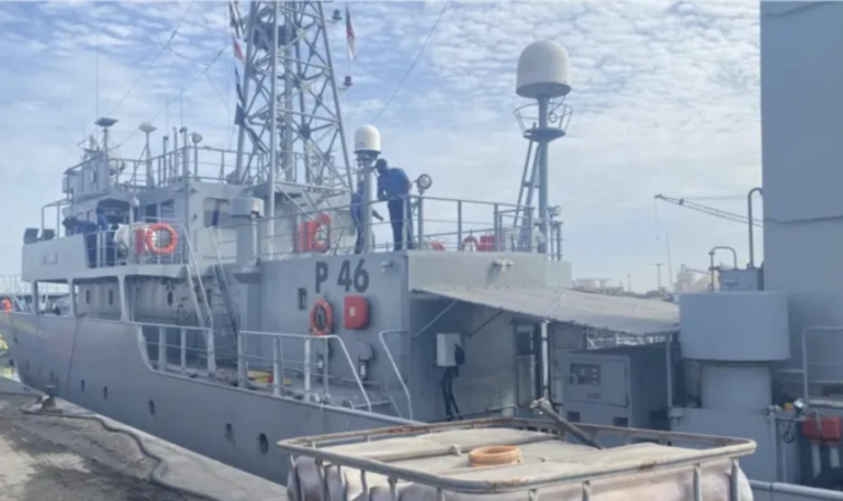 Grey patrol/research vessel docked at a pier, with crew on the upper deck near antennas and radar domes.
