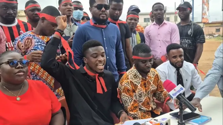 Group of people at an outdoor press conference; a man in black raises his fist with a red headband while reporters hold microphones nearby.