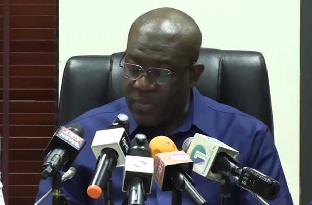 Man in a blue shirt speaks at a press conference, seated behind a row of multiple microphones.