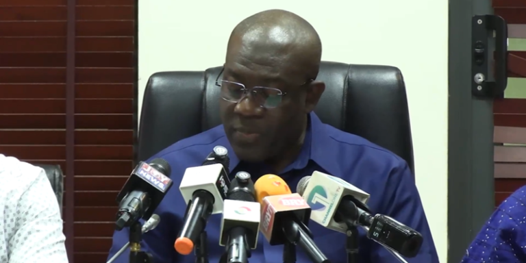 Man in a blue shirt speaks at a press conference, seated behind a row of multiple microphones.
