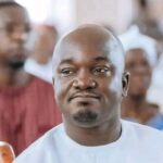 Man in white traditional attire seated at a crowded event, looking slightly to the right with a neutral expression.