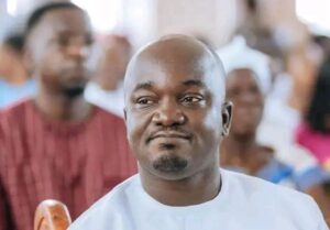 Man in white traditional attire seated at a crowded event, looking slightly to the right with a neutral expression.