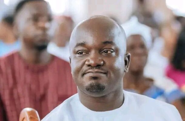 Man in white traditional attire seated at a crowded event, looking slightly to the right with a neutral expression.