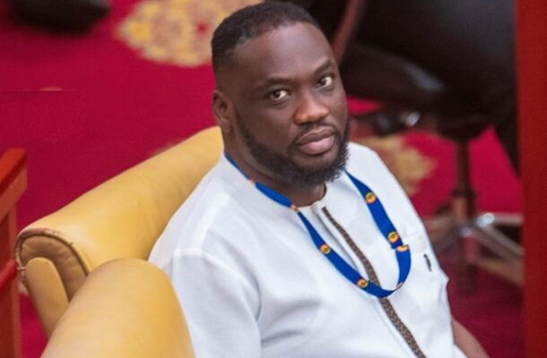 Man in white traditional attire sits on a yellow sofa at an indoor event, wearing a blue lanyard around his neck.