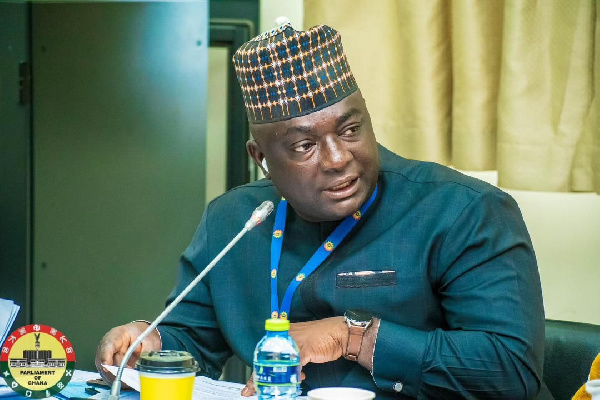 Man wearing a patterned cap speaks into a microphone at a conference table during a formal meeting, with a blue lanyard and water bottle nearby.