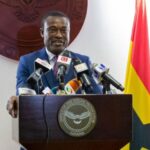 Ghanaian official in a blue suit speaks at a podium with multiple microphones, behind a seal reading 'Office of the Special Prosecutor, Ghana' and a Ghana flag in the background.