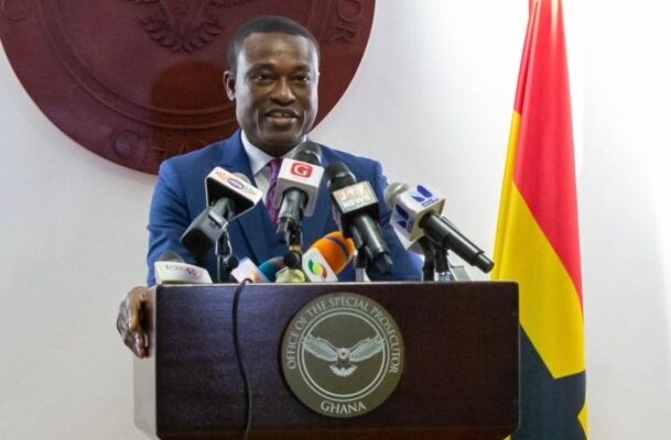 Ghanaian official in a blue suit speaks at a podium with multiple microphones, behind a seal reading 'Office of the Special Prosecutor, Ghana' and a Ghana flag in the background.
