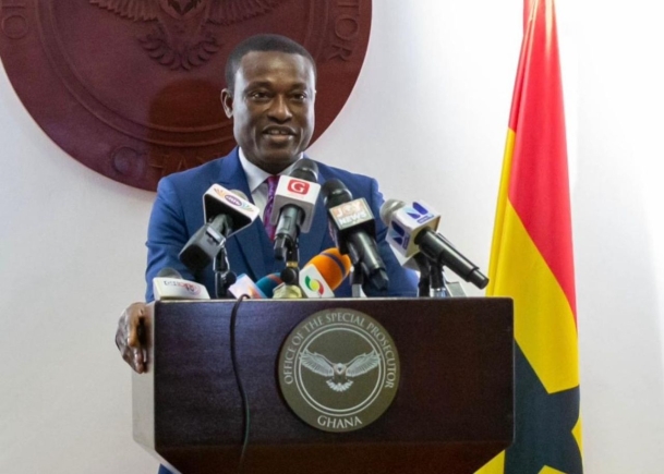 Ghanaian official in a blue suit speaks at a podium with multiple microphones, behind a seal reading 'Office of the Special Prosecutor, Ghana' and a Ghana flag in the background.