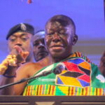 Man wearing colorful traditional fabric speaks at a podium with a microphone, flanked by others at a ceremony.