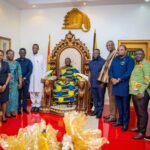 Group of officials posing with a seated dignitary on an ornate throne in a ceremonial room, Ghanaian flags in the background and a crest on the wall.