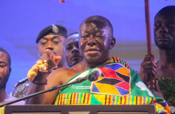 Man wearing colorful traditional fabric speaks at a podium with a microphone, flanked by others at a ceremony.