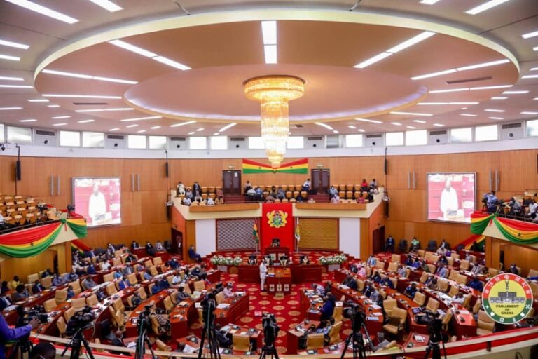 Wide view of a parliament chamber with a circular ceiling chandelier, Ghana flag, and lawmakers seated in red and tan benches around the dais.