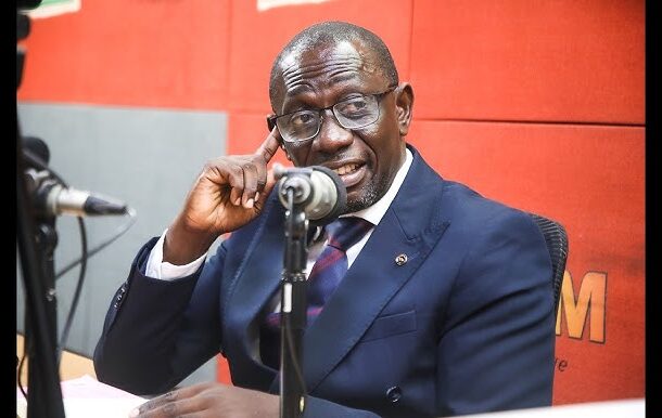 Man in a navy suit and glasses speaks into a desktop microphone during a radio interview on a red-background set.