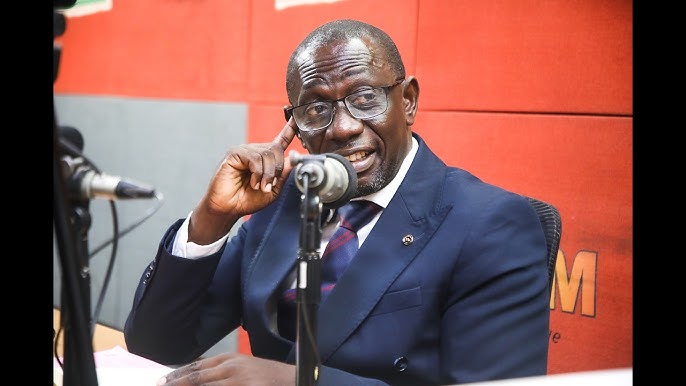 Man in a navy suit and glasses speaks into a desktop microphone during a radio interview on a red-background set.