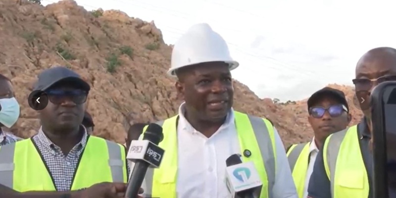 Man in a white hard hat and high-visibility vest speaks to reporters at a construction site with a dirt mound in the background.