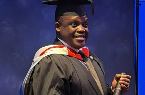 Male graduate in a cap and gown smiling on stage while holding a rolled diploma.