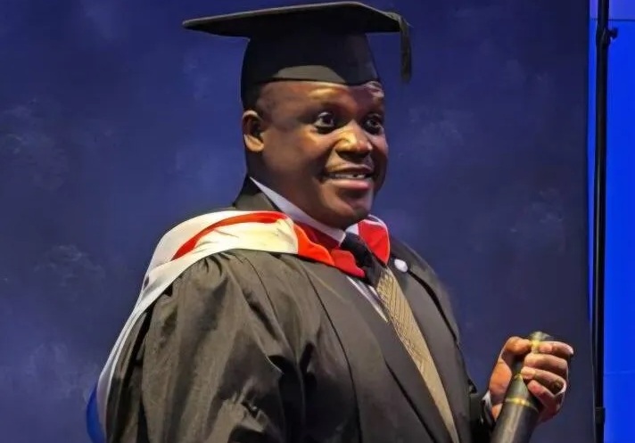 Male graduate in a cap and gown smiling on stage while holding a rolled diploma.