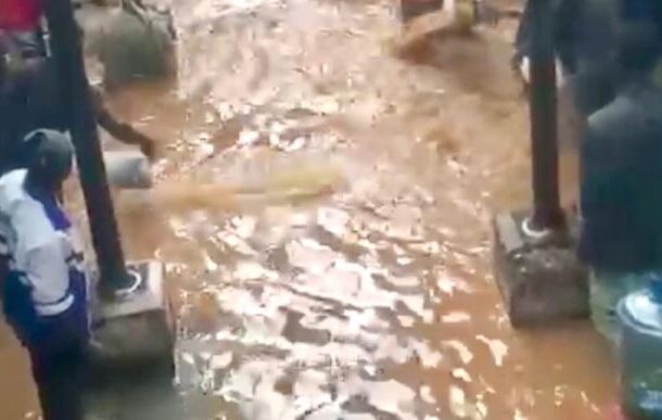 A muddy, waterlogged street with brown floodwater and people standing on concrete blocks along the edges.