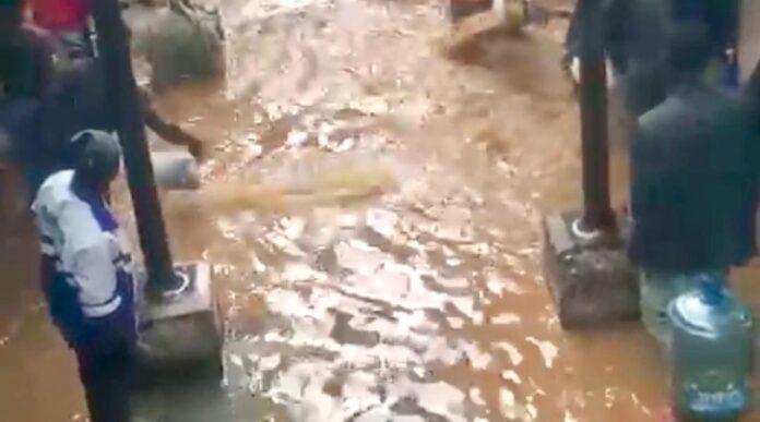 A muddy, waterlogged street with brown floodwater and people standing on concrete blocks along the edges.