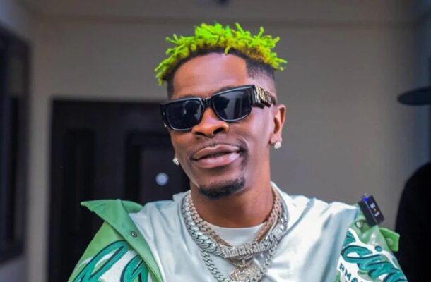 Man with neon green hair, dark sunglasses, and diamond chains posing in a doorway outside a building.