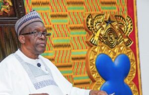 Older man in white traditional attire and a patterned cap, seated beside a colorful woven backdrop and a gold crest with a blue heart.
